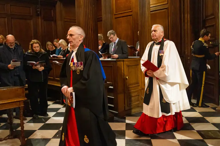 Two clergy members in ceremonial robes and medals walk through a wood-panelled chapel with a checkered floor.