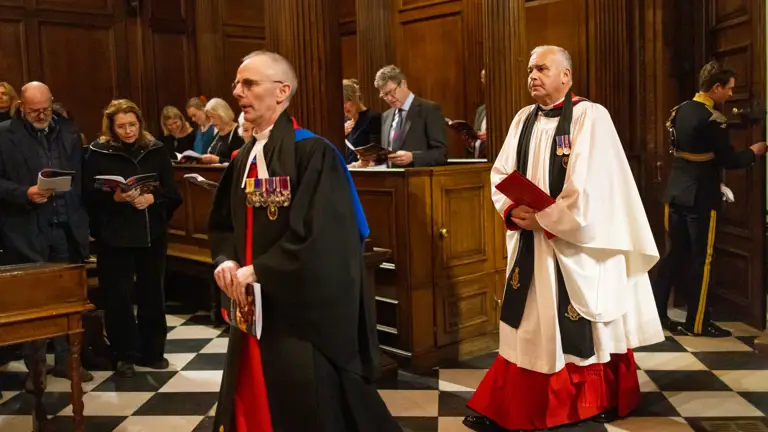 Two clergy members in ceremonial robes and medals walk through a wood-panelled chapel with a checkered floor.