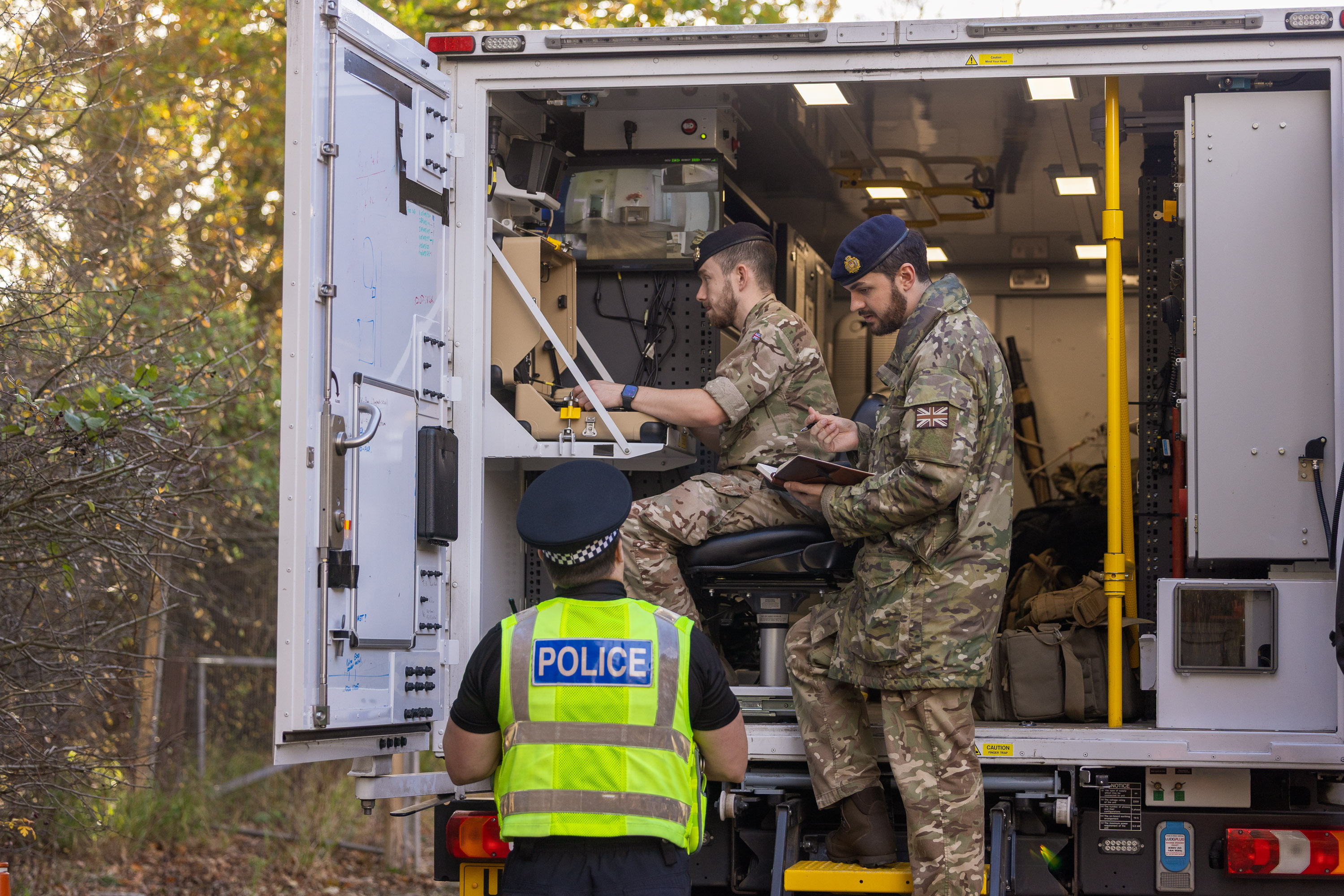 Soldiers in camouflage uniforms sit in the back of a white van while speaking to a police officer wearing a hi-vis vest.