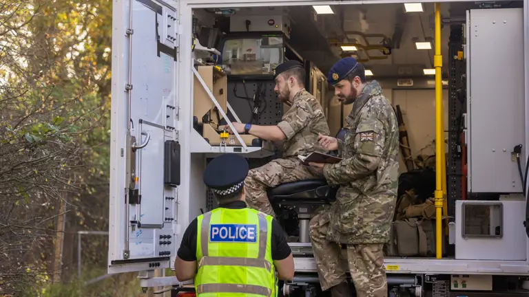 Soldiers in camouflage uniforms sit in the back of a white van while speaking to a police officer wearing a hi-vis vest.