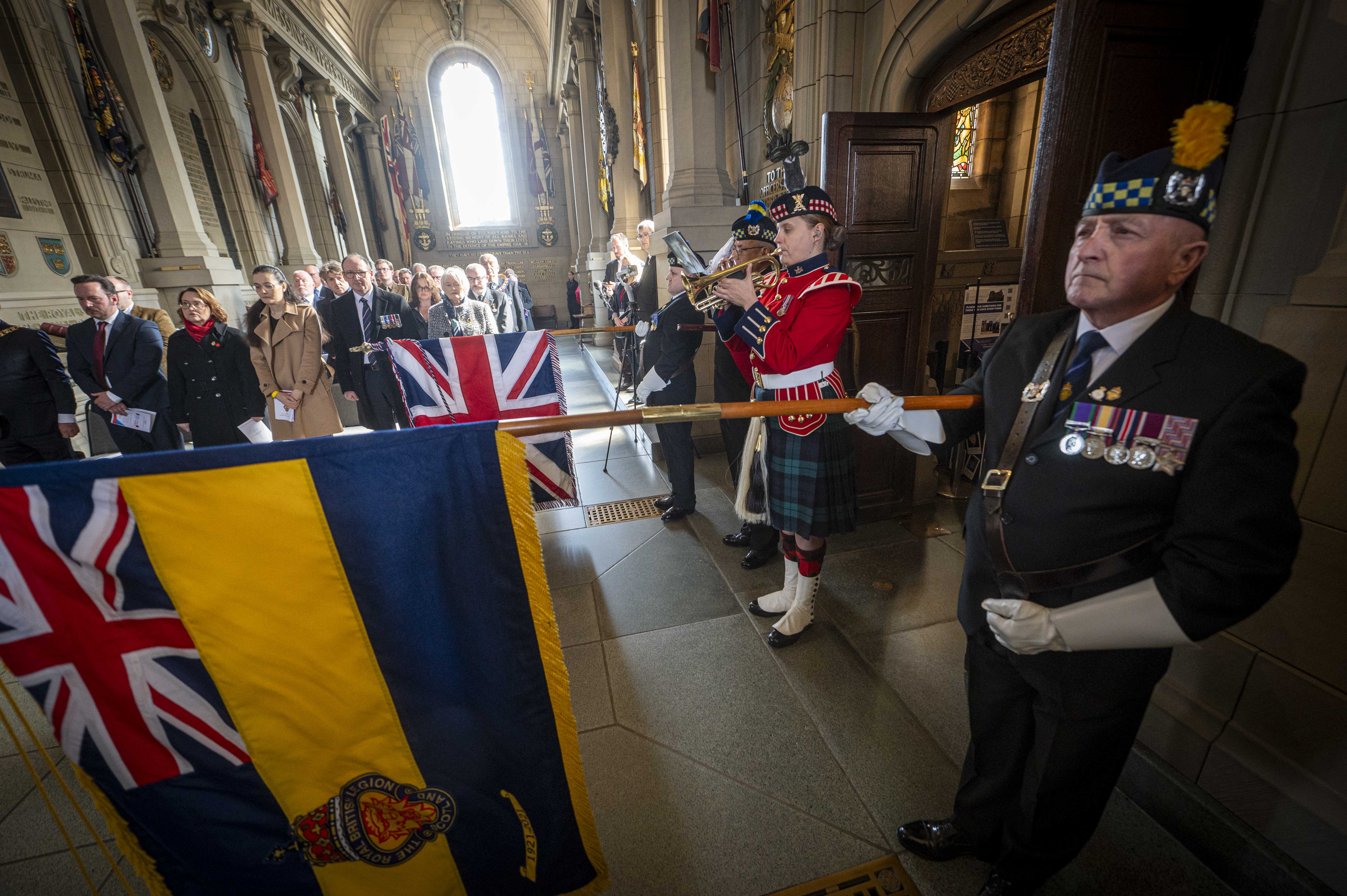 A bugler wearing a red tunic and green kilt plays the bugle. Either side of her, people are lowering flags.
