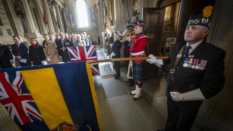 A bugler wearing a red tunic and green kilt plays the bugle. Either side of her, people are lowering flags.