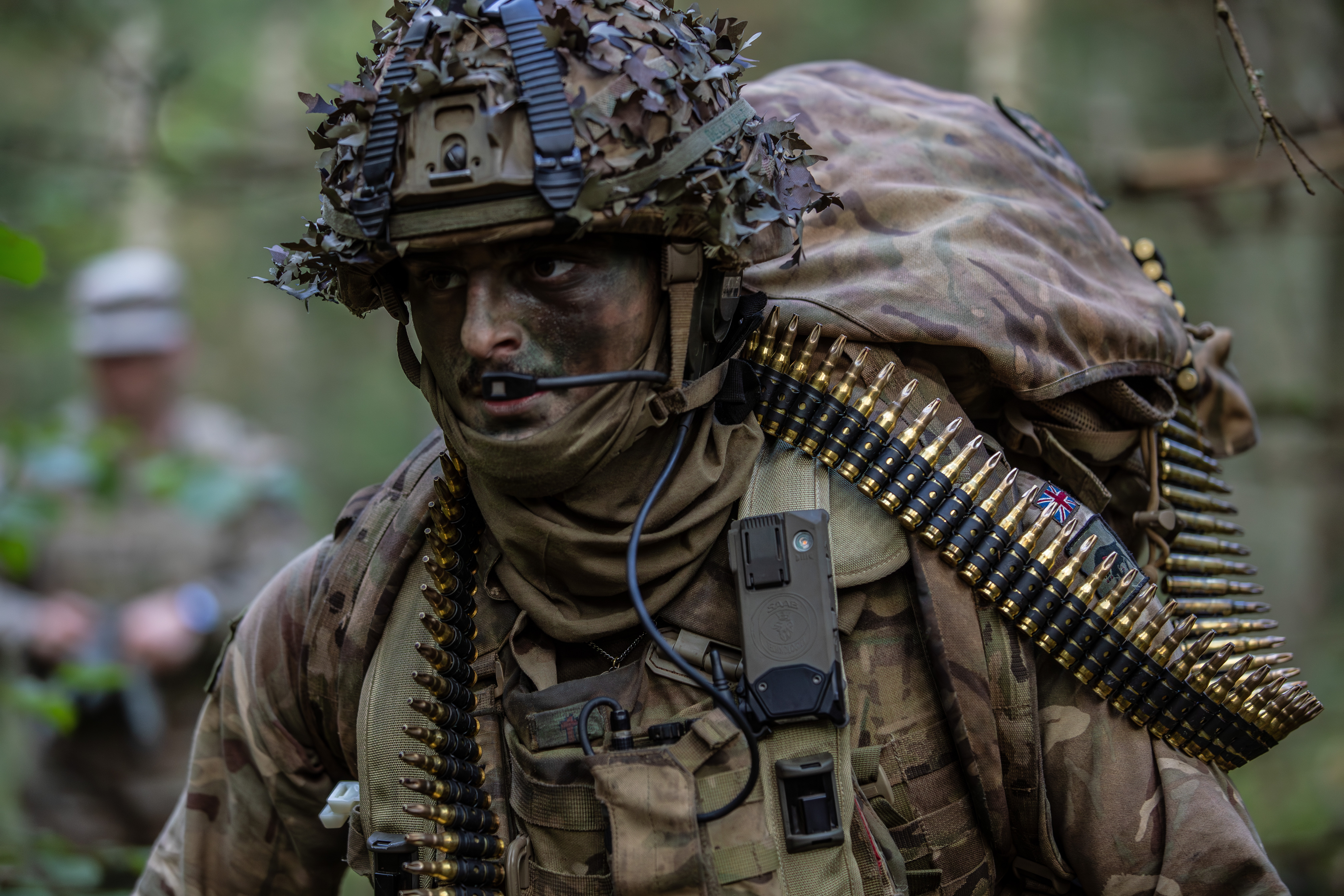 Soldier in camouflage gear with a helmet covered in foliage, carrying ammunition belts and radio equipment.