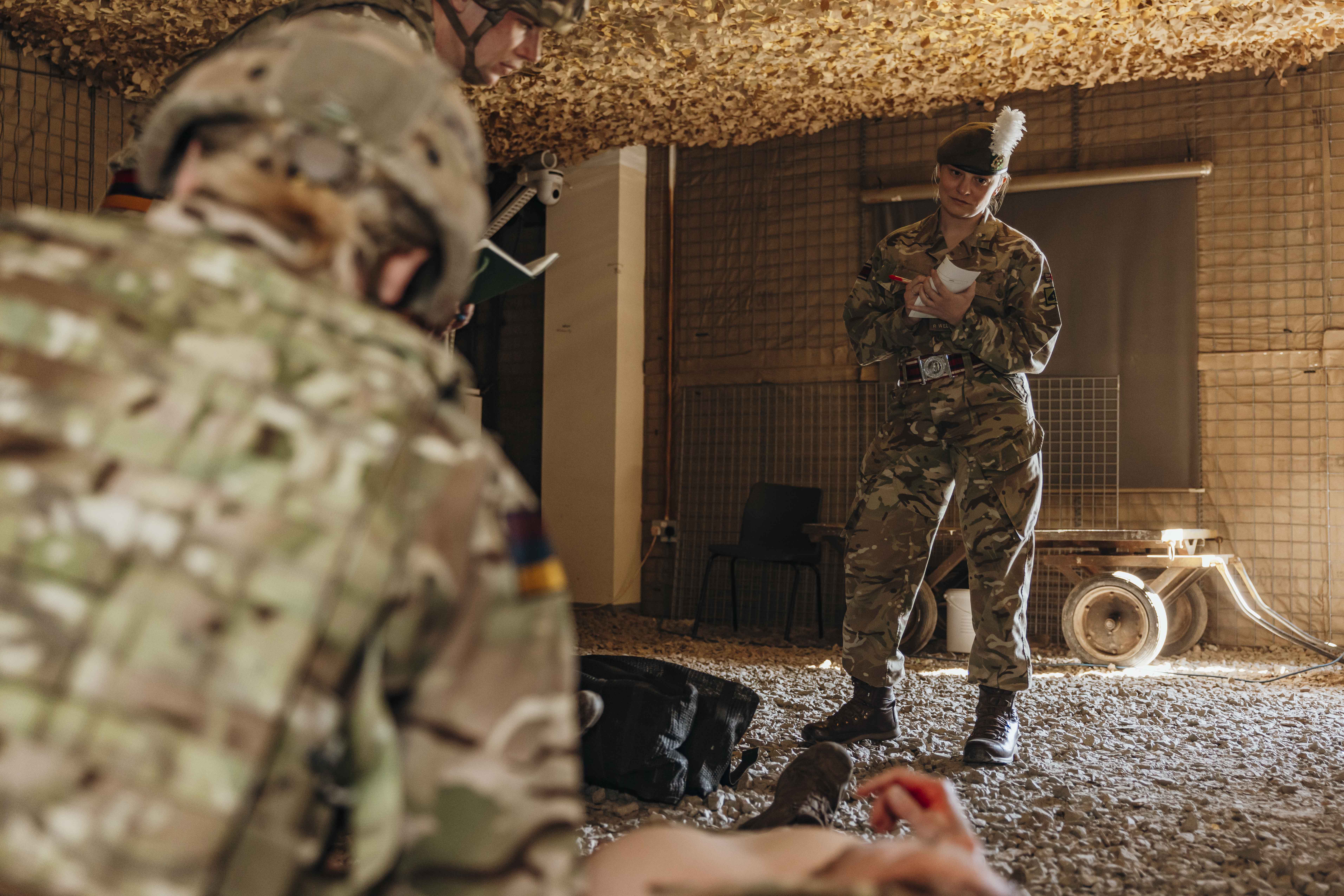 Soldier in camouflage uniform standing inside a makeshift shelter, taking notes on a clipboard during a training exercise.