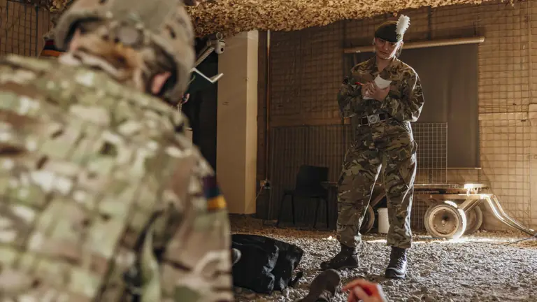 Soldier in camouflage uniform standing inside a makeshift shelter, taking notes on a clipboard during a training exercise.