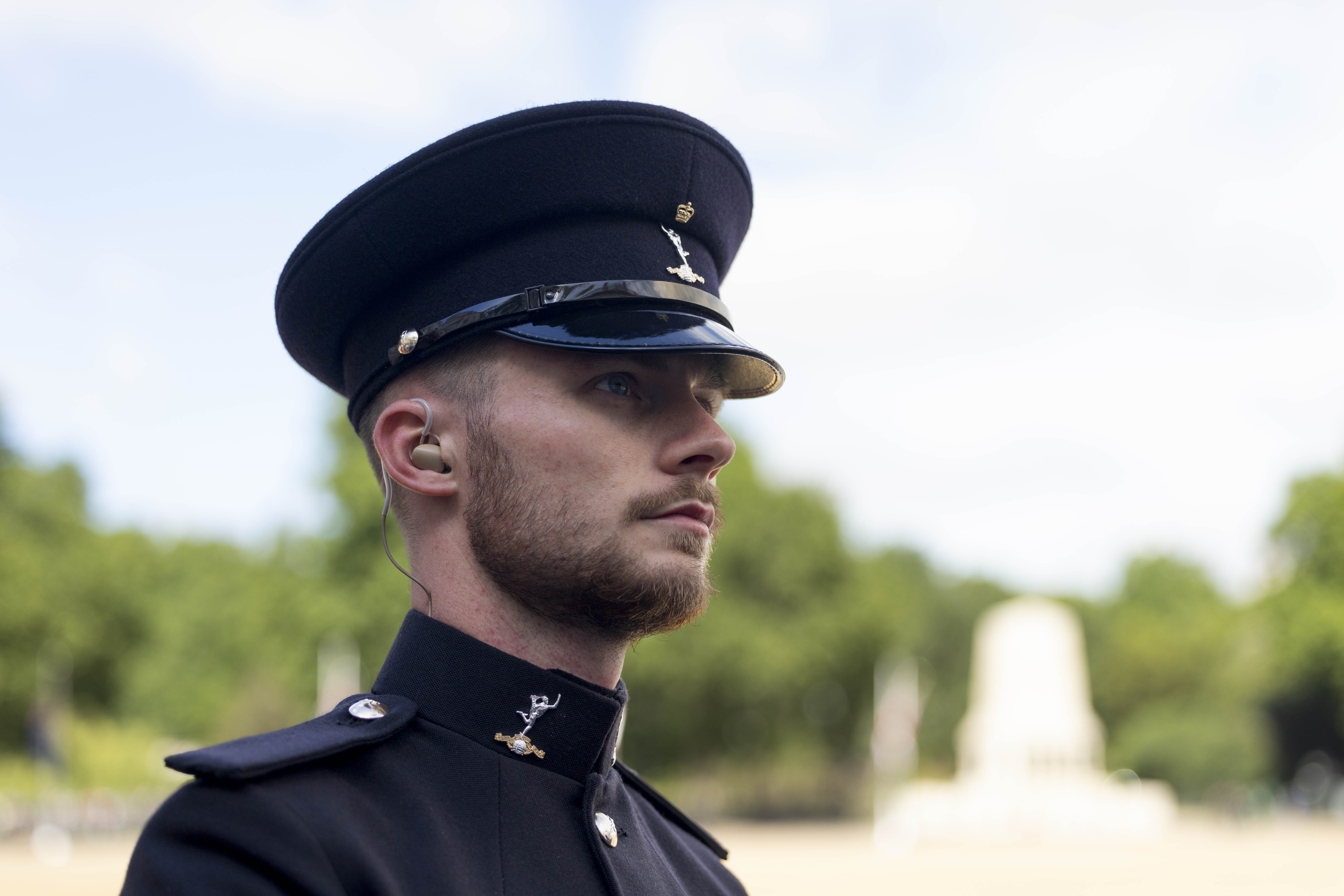 A uniformed man in a black hat and coat stands outside, wearing an earpiece. The background is a park with blurred trees and a monument, conveying formality.
