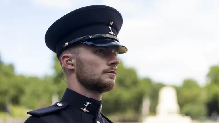 A uniformed man in a black hat and coat stands outside, wearing an earpiece. The background is a park with blurred trees and a monument, conveying formality.