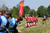 A group of individuals is engaged in a tug of war on a grassy field. One team is wearing red shirts and is pulling the rope towards a banner that reads