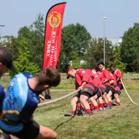 A group of individuals is engaged in a tug of war on a grassy field. One team is wearing red shirts and is pulling the rope towards a banner that reads