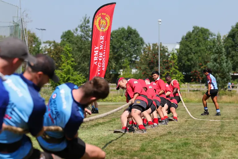 A group of individuals is engaged in a tug of war on a grassy field. One team is wearing red shirts and is pulling the rope towards a banner that reads