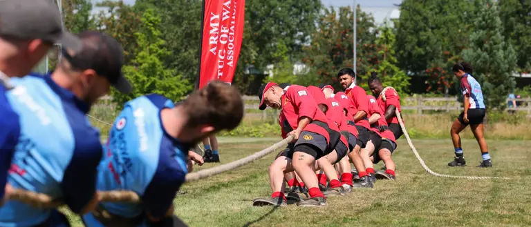 A group of individuals is engaged in a tug of war on a grassy field. One team is wearing red shirts and is pulling the rope towards a banner that reads