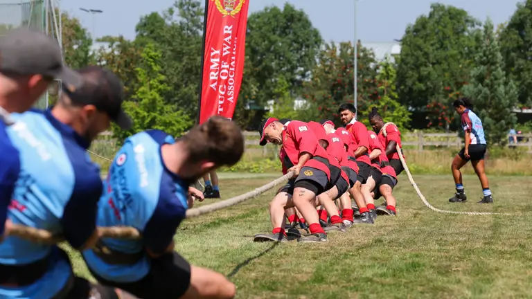 A group of individuals is engaged in a tug of war on a grassy field. One team is wearing red shirts and is pulling the rope towards a banner that reads