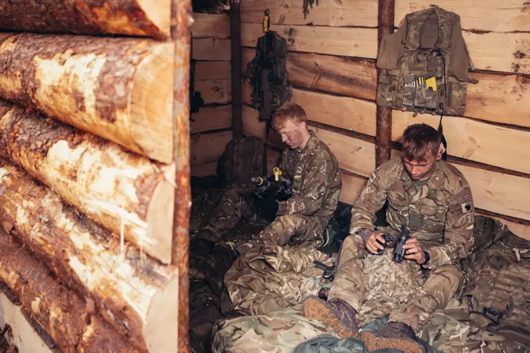 Two soldiers wearing camouflage uniforms sit inside of a wooden shelter which they have built.