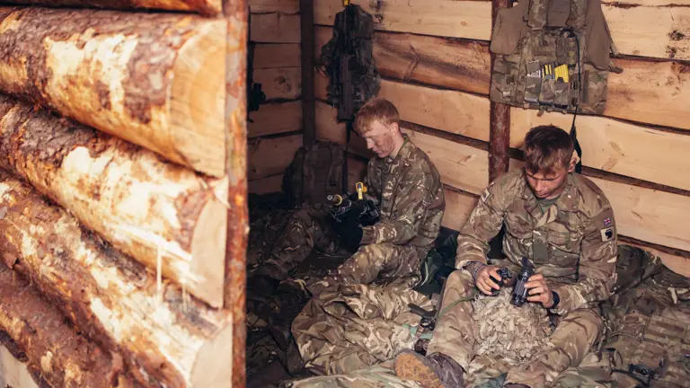 Two soldiers wearing camouflage uniforms sit inside of a wooden shelter which they have built.