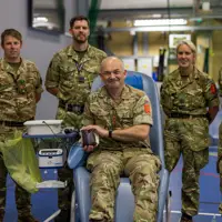 Group of five soldiers in camouflage uniforms inside a gymnasium, one seated donating blood.