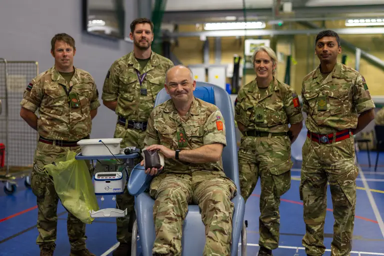 Group of five soldiers in camouflage uniforms inside a gymnasium, one seated donating blood.