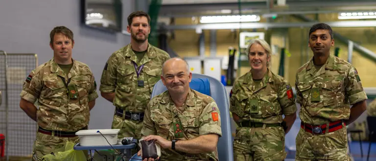 Group of five soldiers in camouflage uniforms inside a gymnasium, one seated donating blood.
