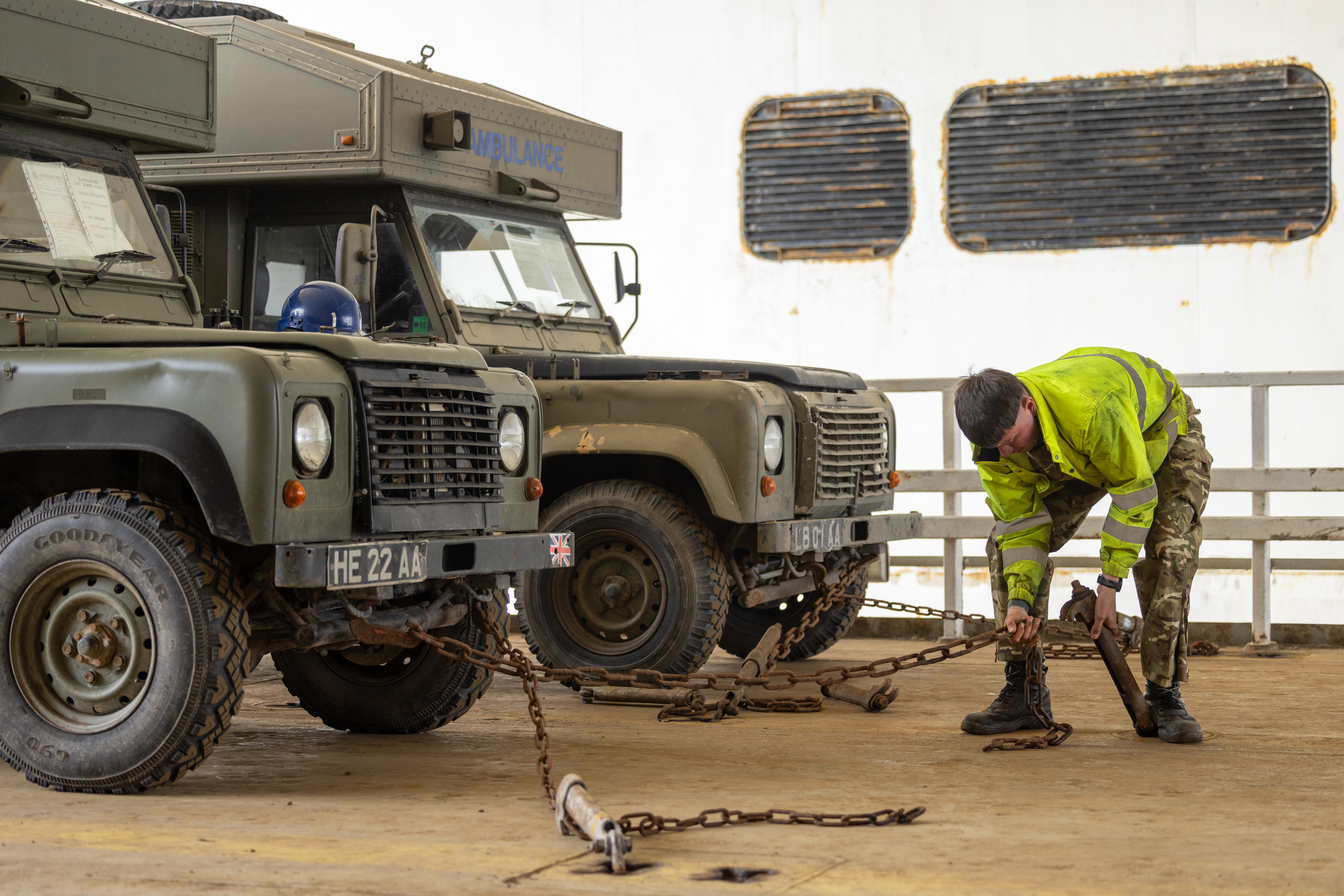 Military personnel in camouflage and high-visibility jacket securing chains to two green vehicles.