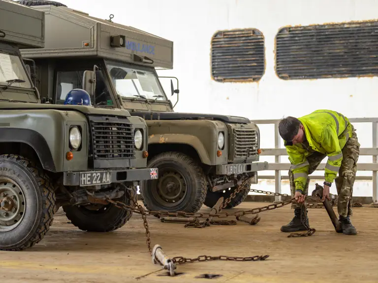 Military personnel in camouflage and high-visibility jacket securing chains to two green vehicles.