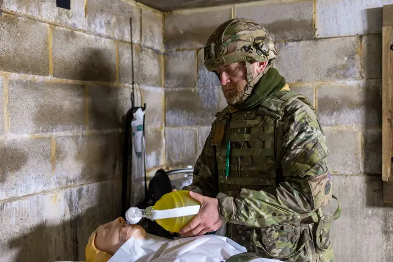 Soldier in camouflage uniform practicing emergency ventilation on a medical training mannequin inside a concrete room.