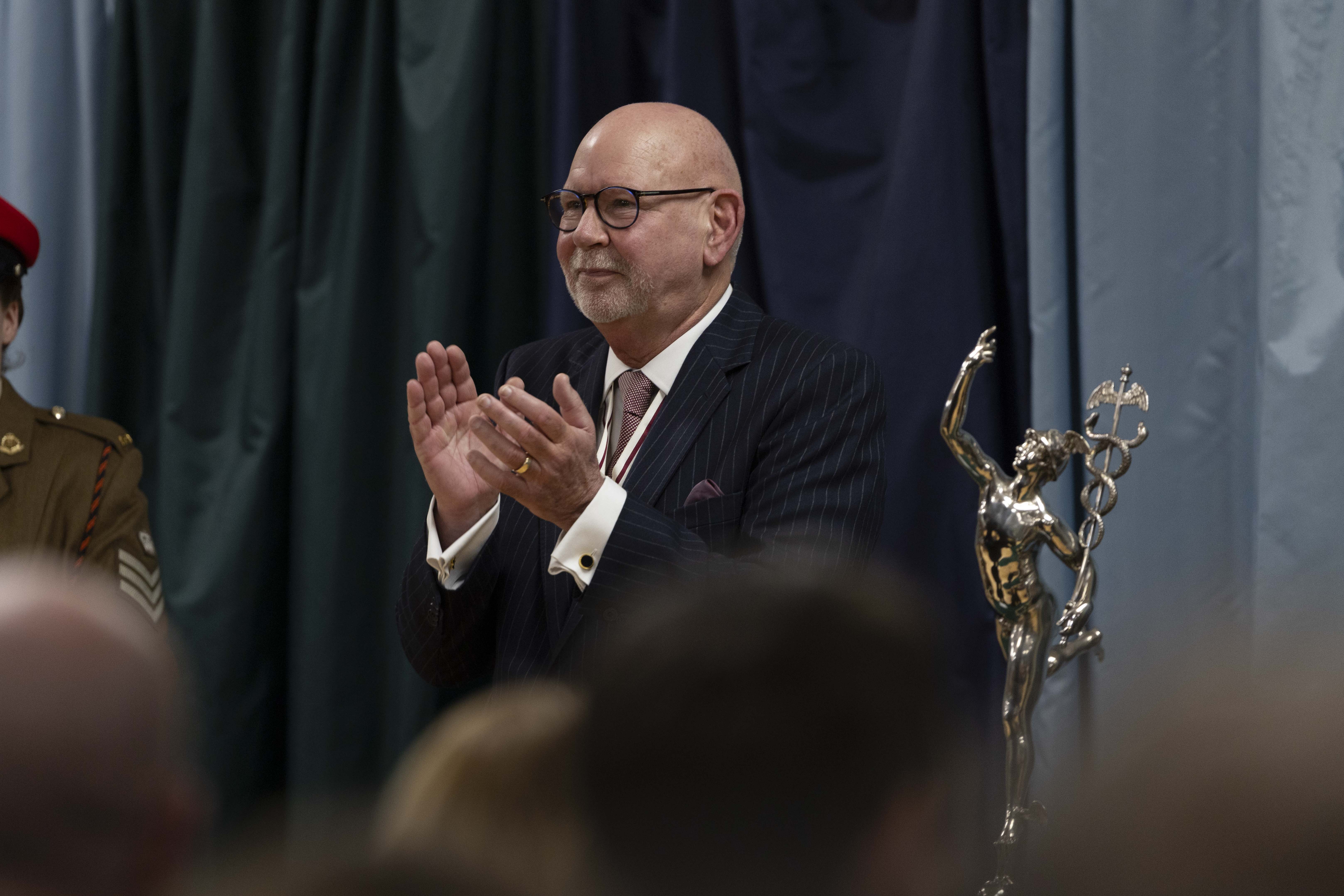 An older man in a suit claps at an event, standing next to a silver statue depicting a winged figure. The background features dark curtains.