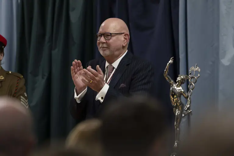 An older man in a suit claps at an event, standing next to a silver statue depicting a winged figure. The background features dark curtains.