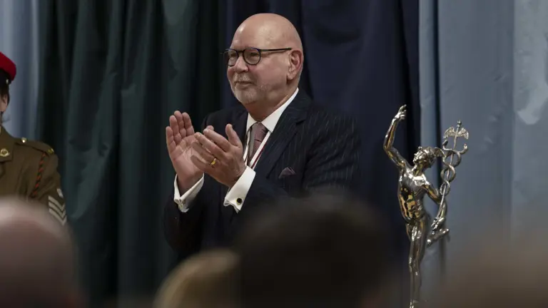 An older man in a suit claps at an event, standing next to a silver statue depicting a winged figure. The background features dark curtains.