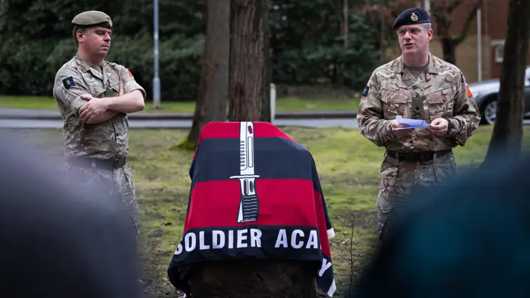Two senior Army personnel in uniform stand either side of a black and red striped flag.