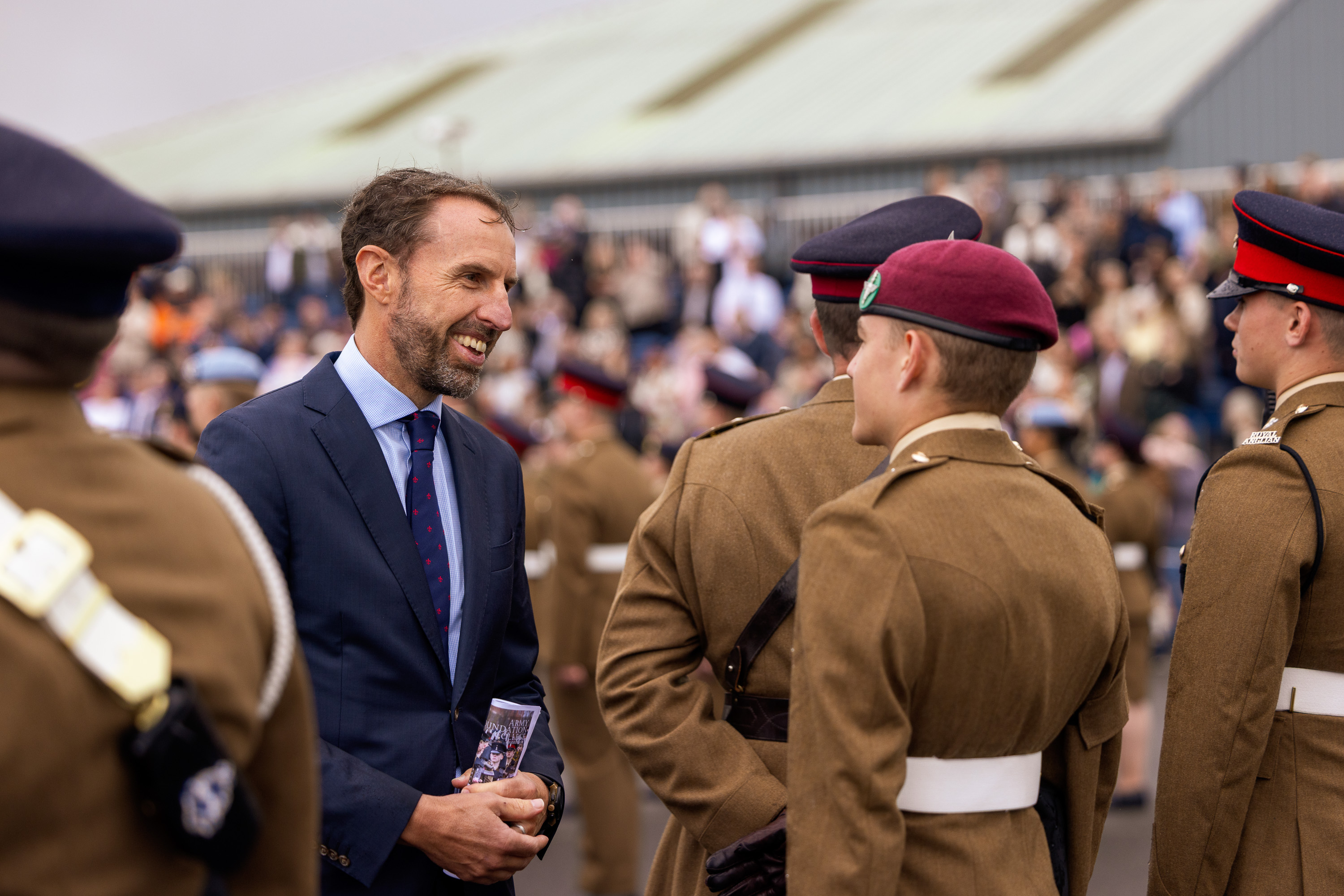 Soldiers stood for inspection are seen chatting with Sir Gareth Southgate who is wearing a suit and tie. Family and friends are seen in the crowd in the background.