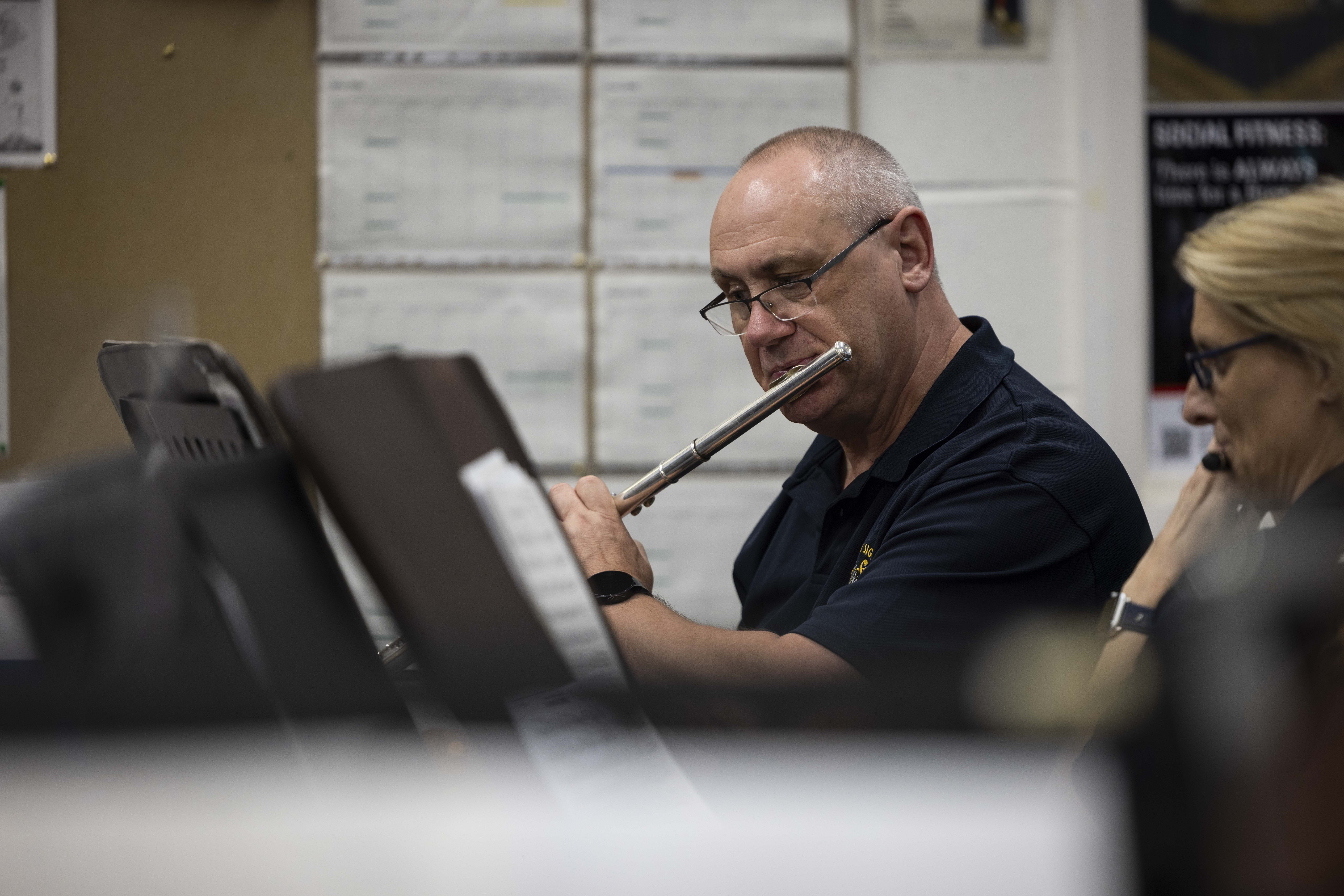 A person is playing the flute in a music ensemble setting. They're seated amongst others surrounded by music stands. 