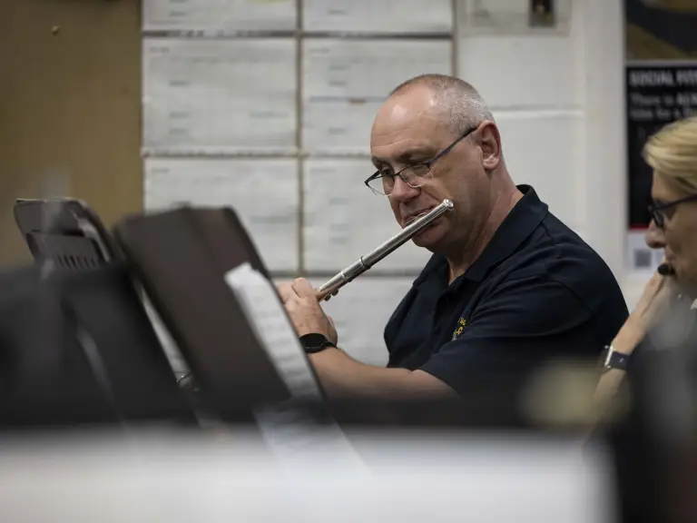 A person is playing the flute in a music ensemble setting. They're seated amongst others surrounded by music stands.