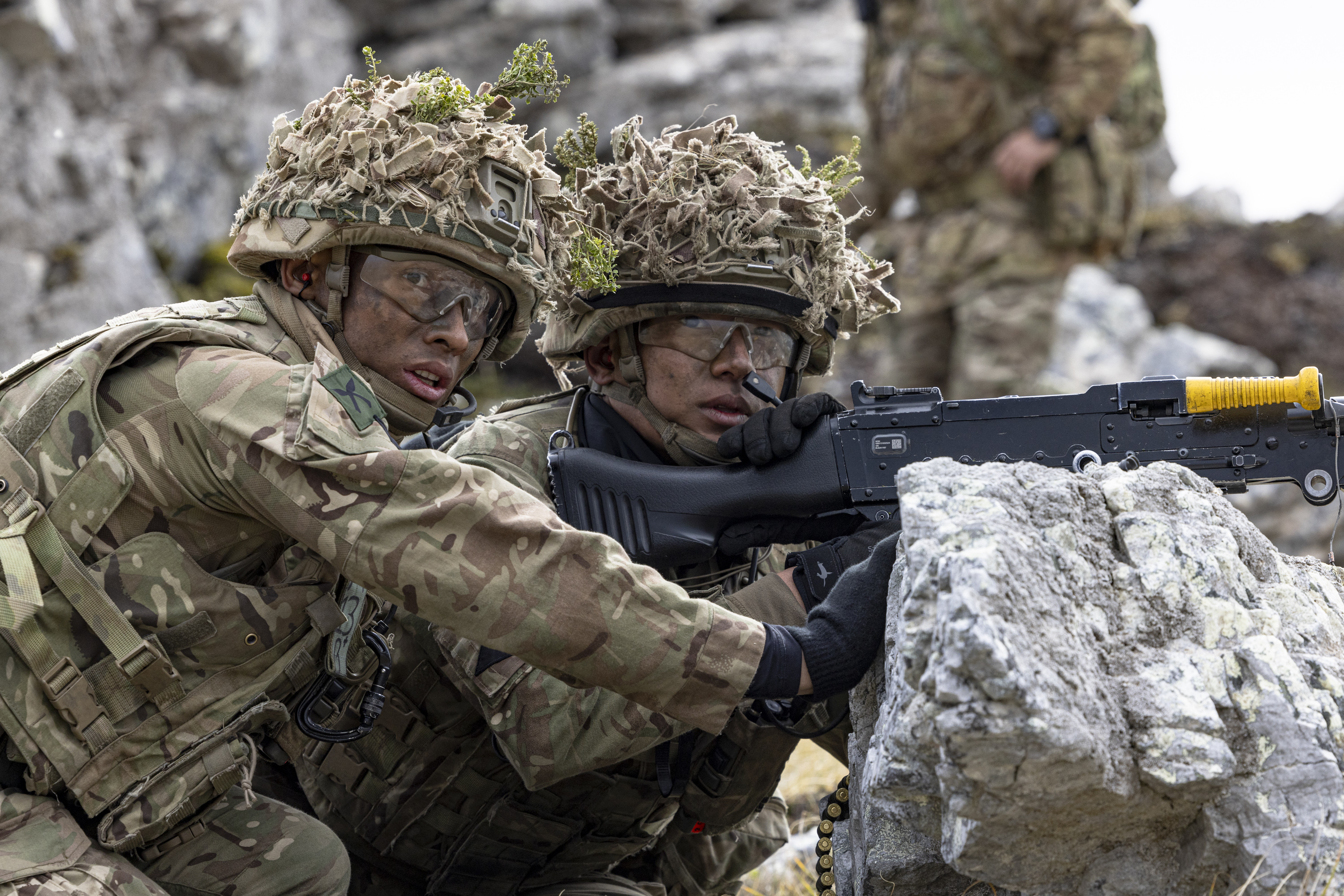 Two soldiers in camouflage uniforms, wearing helmets with foliage, are positioned behind rocks.