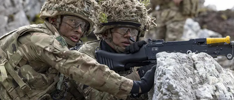 Two soldiers in camouflage uniforms, wearing helmets with foliage, are positioned behind rocks.