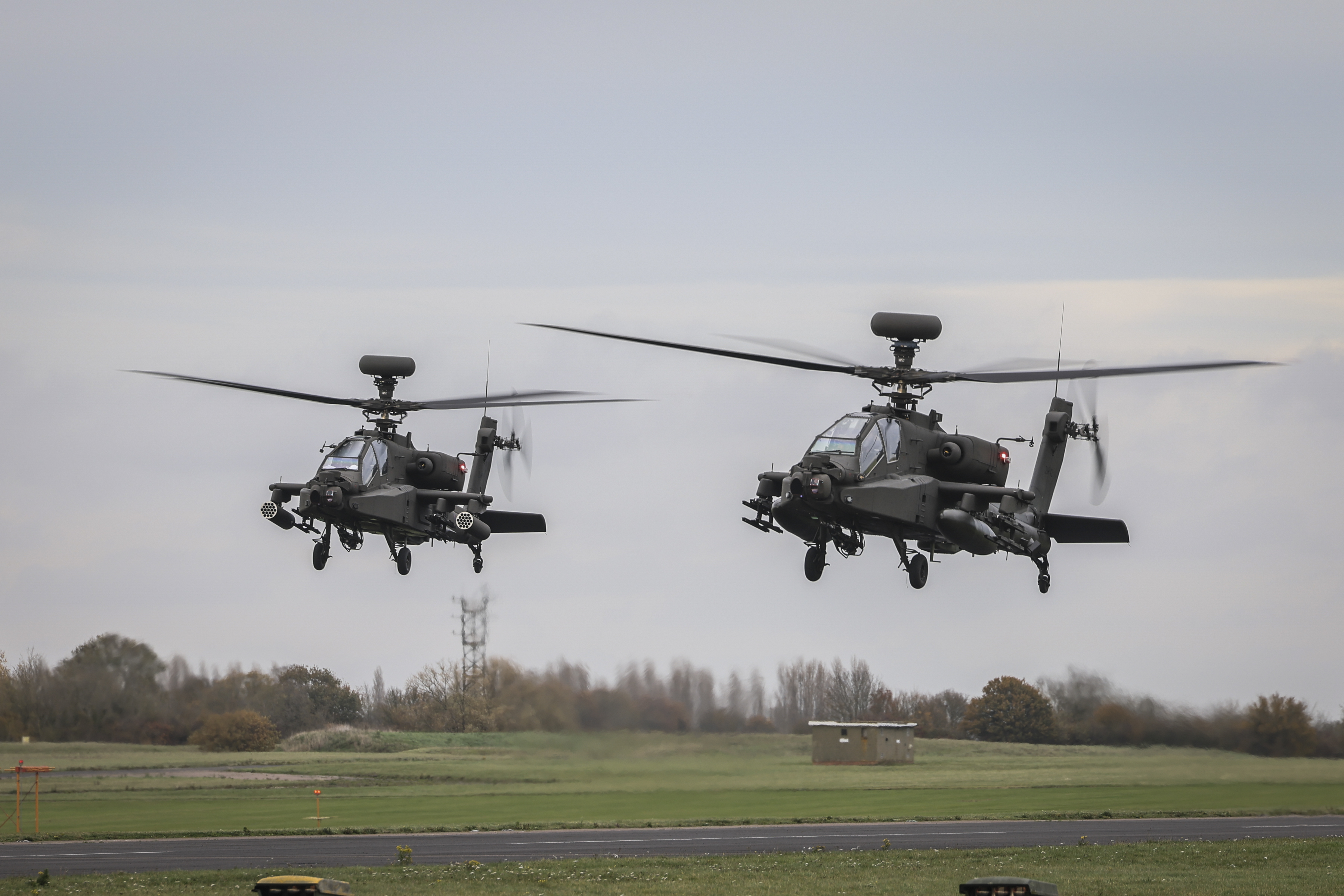 Two Apache helicopters hover above a grassy field with a clear sky in the background.