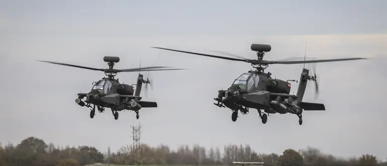 Two Apache helicopters hover above a grassy field with a clear sky in the background.