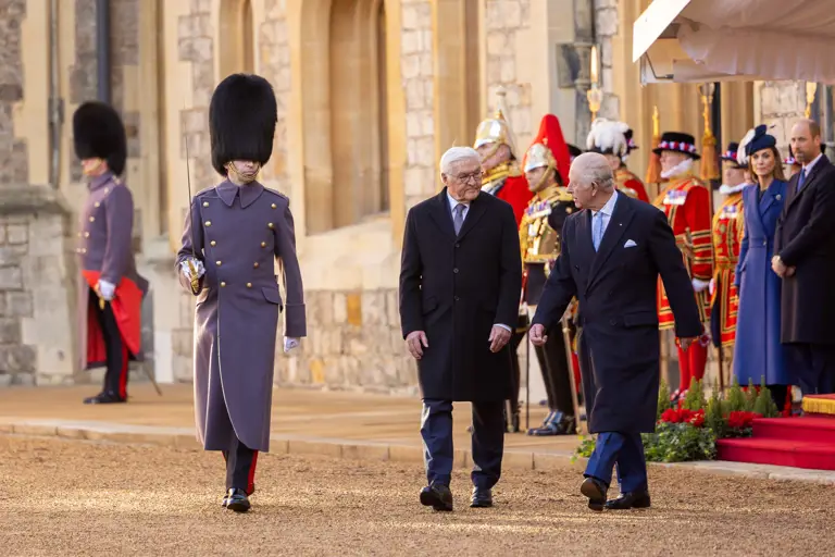 A formal procession at a historic building. A guard in a tall black hat leads two elderly men in overcoats. Onlookers and more guards in ceremonial attire are in the background.