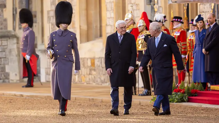 A formal procession at a historic building. A guard in a tall black hat leads two elderly men in overcoats. Onlookers and more guards in ceremonial attire are in the background.