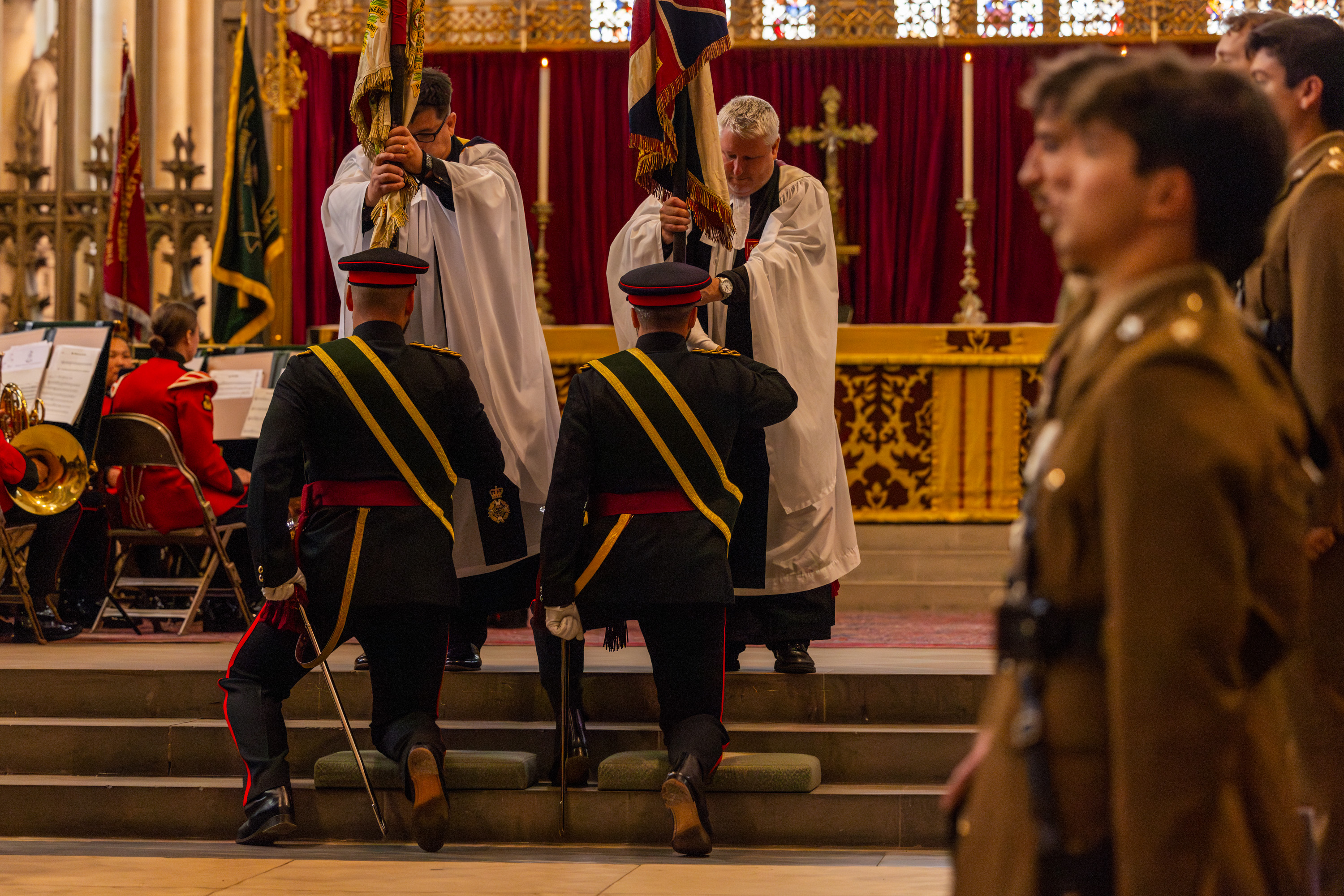 A ceremonial event inside a church features individuals in uniforms and robes. Two uniformed individuals kneel, holding flags while two others in robes stand close by. A band is visible seated on the left, and a large cross is on display in the background. The scene reflects a formal military or religious ceremony.