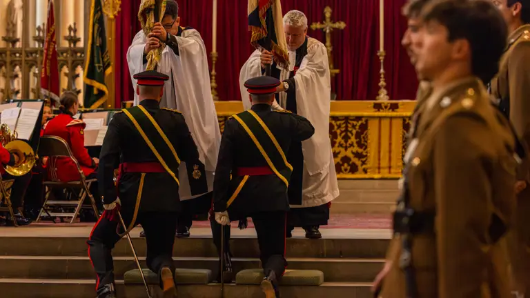 A ceremonial event inside a church features individuals in uniforms and robes. Two uniformed individuals kneel, holding flags while two others in robes stand close by. A band is visible seated on the left, and a large cross is on display in the background. The scene reflects a formal military or religious ceremony.