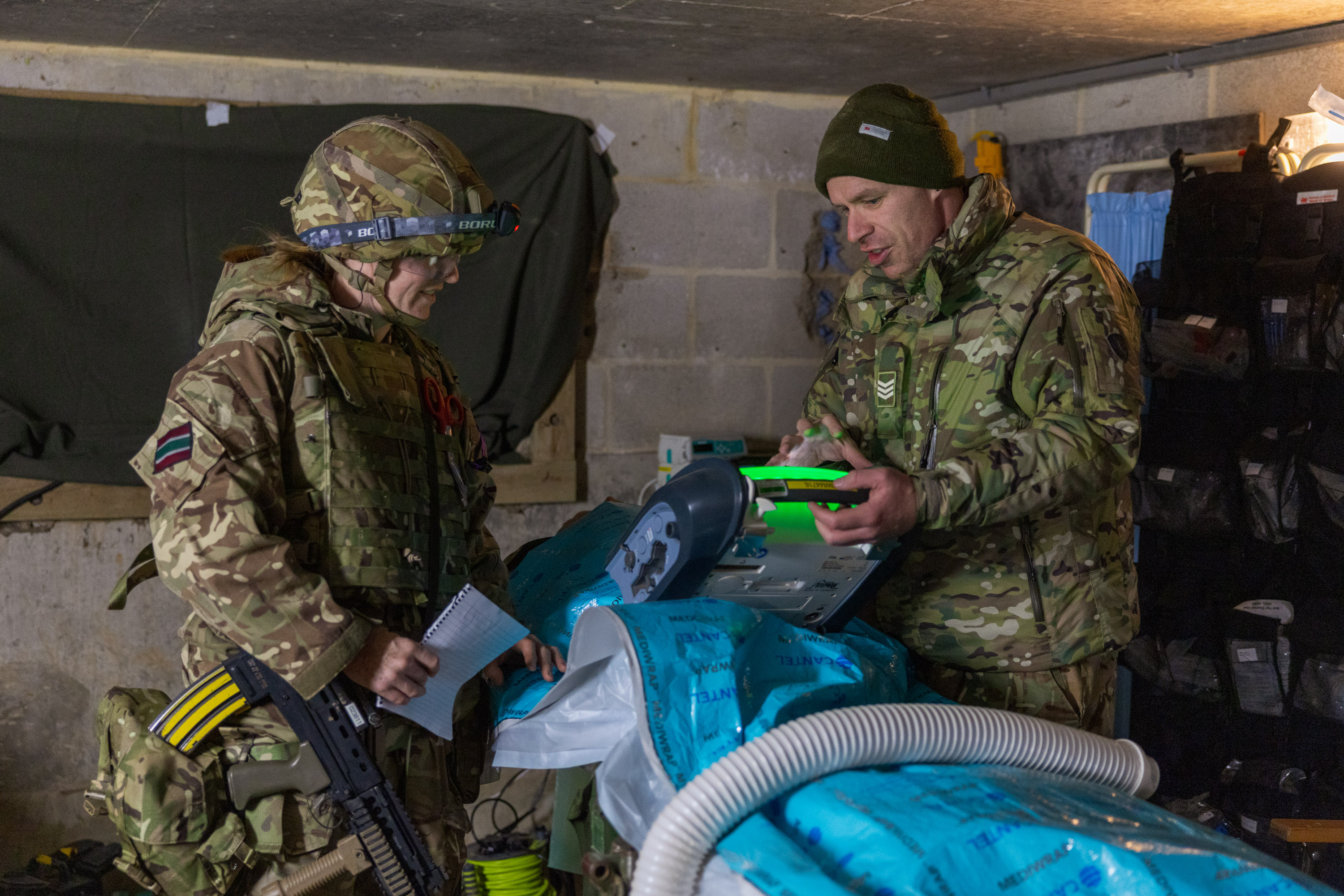 Two soldiers in camouflage gear operate medical equipment inside a concrete bunker room with supplies.