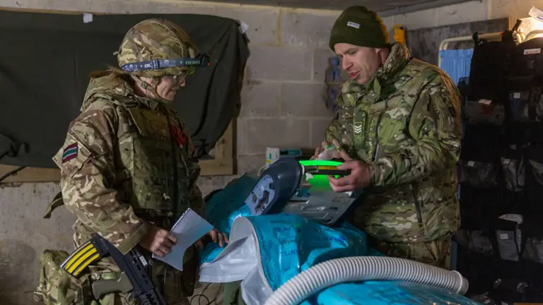 Two soldiers in camouflage gear operate medical equipment inside a concrete bunker room with supplies.