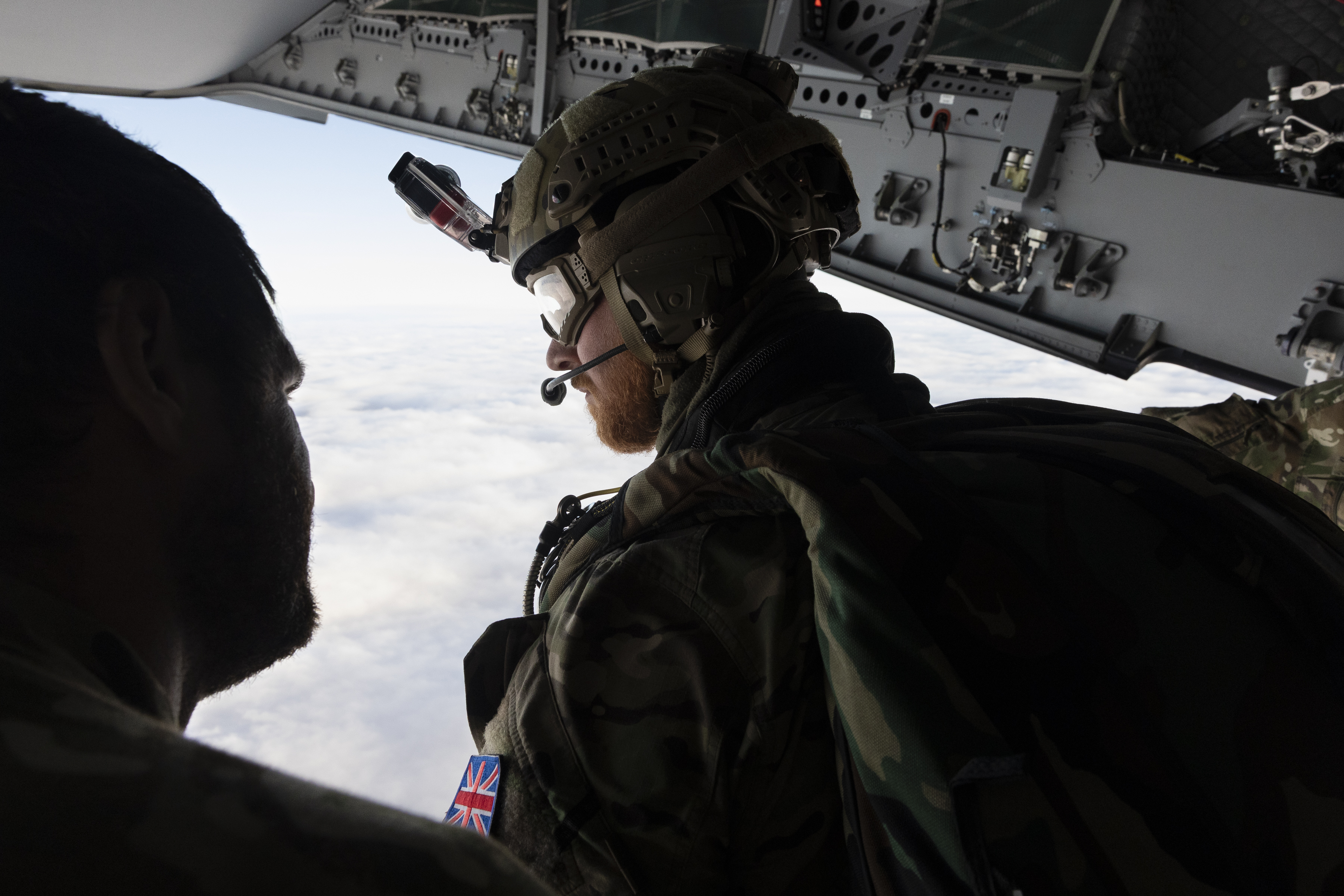Two individuals in military gear are inside an aircraft, looking out over a cloudy sky. One person is wearing a helmet with a camera, gear, and patches with the UK flag. The interior of the aircraft shows various equipment and panels.