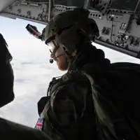 Two individuals in military gear are inside an aircraft, looking out over a cloudy sky. One person is wearing a helmet with a camera, gear, and patches with the UK flag. The interior of the aircraft shows various equipment and panels.