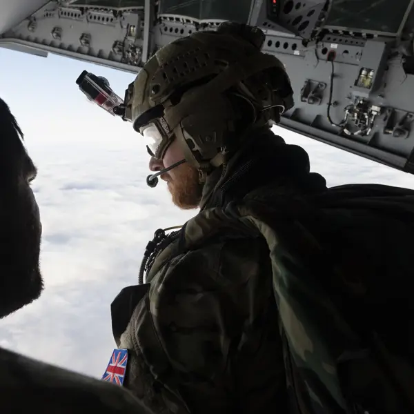 Two individuals in military gear are inside an aircraft, looking out over a cloudy sky. One person is wearing a helmet with a camera, gear, and patches with the UK flag. The interior of the aircraft shows various equipment and panels.