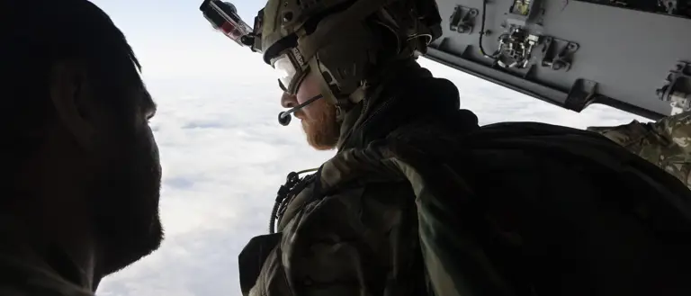 Two individuals in military gear are inside an aircraft, looking out over a cloudy sky. One person is wearing a helmet with a camera, gear, and patches with the UK flag. The interior of the aircraft shows various equipment and panels.