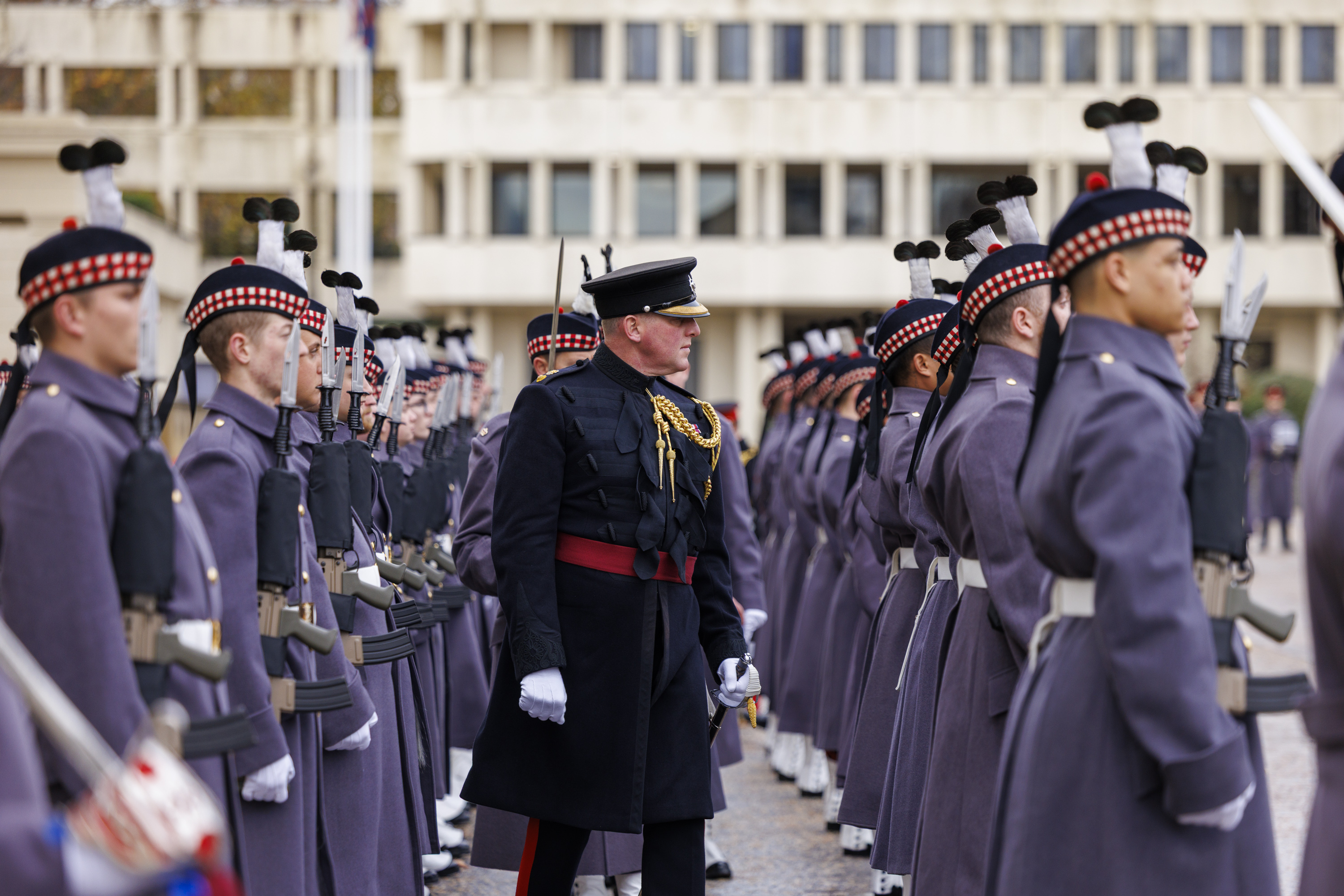 A military officer inspects a line of soldiers dressed in grey uniforms and traditional Scottish hats.