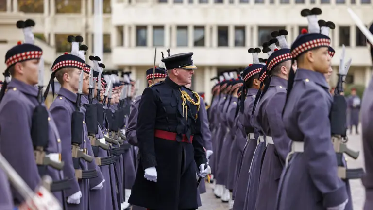 A military officer inspects a line of soldiers dressed in grey uniforms and traditional Scottish hats.