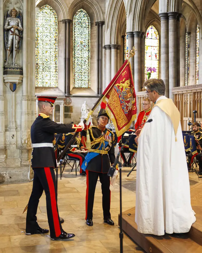 Two military officers in ceremonial uniforms hold a red flag inside a grand cathedral with stained glass windows and stone arches.