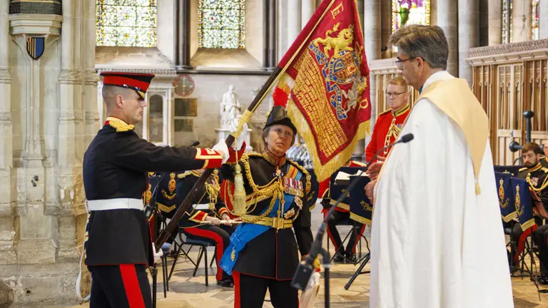 Two military officers in ceremonial uniforms hold a red flag inside a grand cathedral with stained glass windows and stone arches.