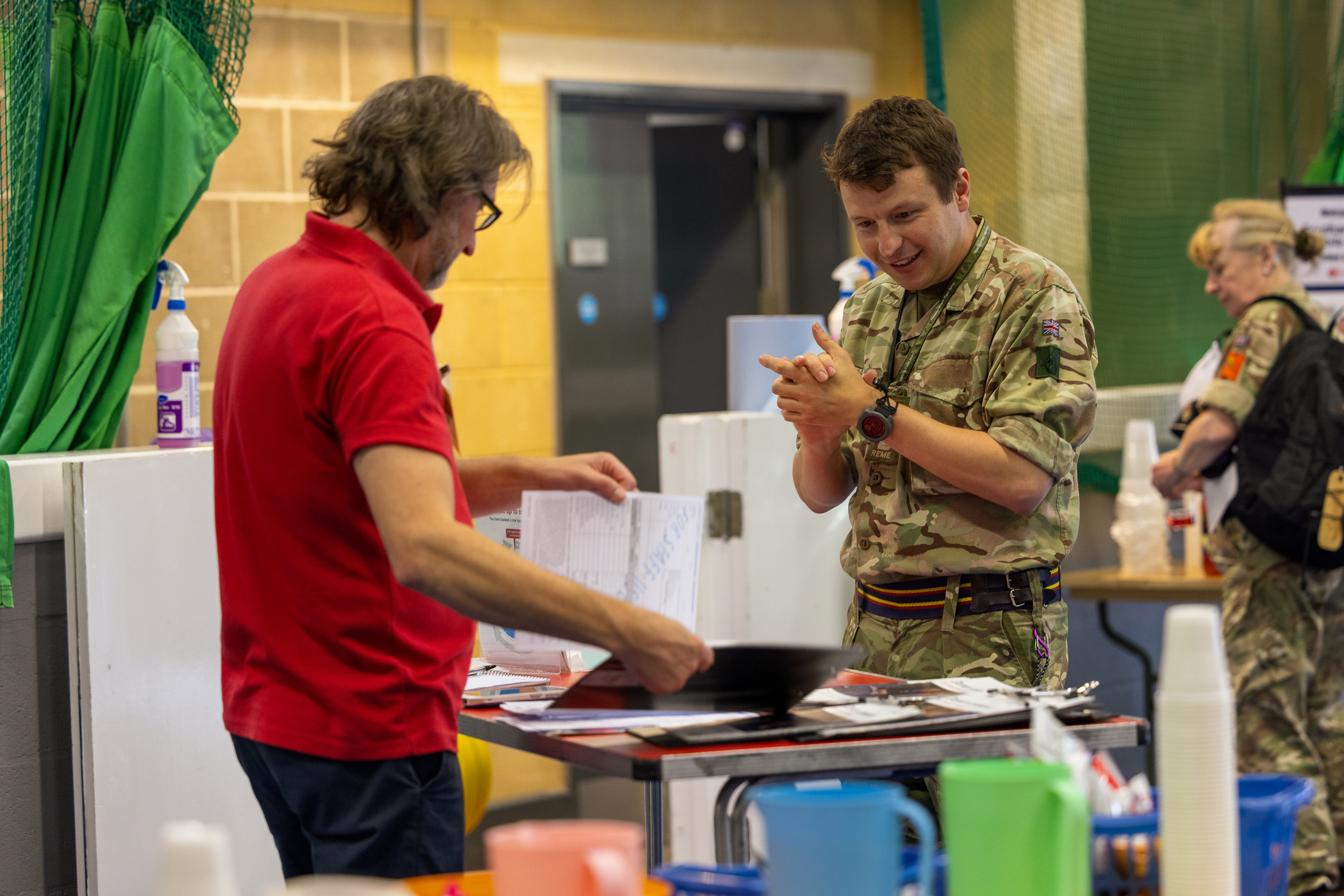 A man in a red shirt reviews documents while a soldier in camouflage listens attentively indoors.